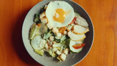 Taze yeşillik, dilimlenmiş tavuk, kızarmış ekmek, kızarmış yumurta ve kiraz domatesli renkli bir salata kasesi ahşap bir masada servis edilir..