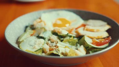 Bir kase taze yeşillik, dilimlenmiş salatalık, domates ve üzeri kızarmış yumurta ve kızarmış tavuk parçaları olan salata. Tabak, ahşap bir masada şık bir kasede servis edilir..