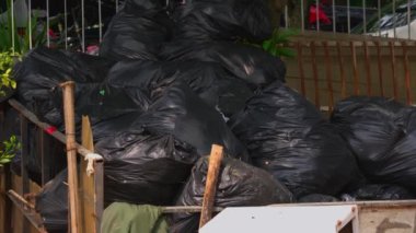 Etrafı yeşilliklerle çevrili bir çite rastgele dizilmiş büyük bir yığın siyah çöp torbası ve arka planda bir anlık da olsa araçlar..