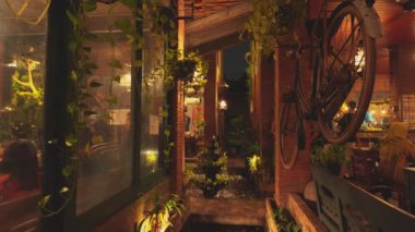 İçinde tuğla duvarlar, asılı bitkiler ve duvara monte edilmiş antika bir bisiklet olan rahat bir kafe. Yumuşak ışık sıcak bir atmosfer yaratıyor ve müşteriler zamanlarının tadını çıkarıyor. Dekor, rustik ve modern elementleri harmanlıyor ve rahat bir hava yaratıyor..