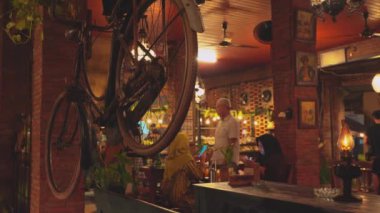 Tavandan sarkan klasik bir bisikletin, sıcak ışıklandırmanın ve yemeklerin keyfini çıkaran müşterilerin olduğu rahat bir restoran içi. Dekorasyonda tuğla duvarlar ve bitkiler var. Hoş bir atmosfer yaratıyorlar..