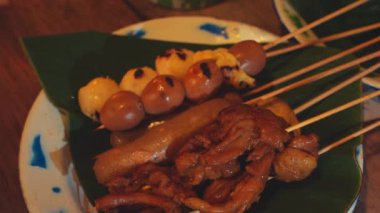 Muz yaprakları üzerinde servis edilen marine edilmiş et ve sebzelerle servis edilen bir tabak ızgara şiş. Tabakta tavuk, sosis ve köfteler yer alıyor, çeşitli doku ve renkler sergileniyor..