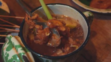 Dekoratif bir tencerede, otlar ve et parçaları içeren geleneksel bir çorbanın yakın çekimi. Çorba buharlanıyor, limon otu ve baharat gibi görünen malzemelerle, kırsal ahşap bir masa üzerine kurulmuş..