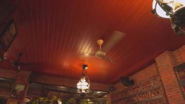Ahşap tavanı kırmızı renge boyanmış samimi bir restoran içi. Bir tavan vantilatörü dönüyor ve dekoratif lambalar tavandan sarkıyor. Sıcak bir ortam yaratıyor..