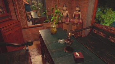 İçinde ahşap mobilyalar, vazolu dekoratif bir masa ve iki ahşap heykel olan geleneksel bir odanın içi. Duvarlar kırmızı tuğlalardan yapılmış ve arka planda sıcak bir atmosfer yaratan bitkiler var..
