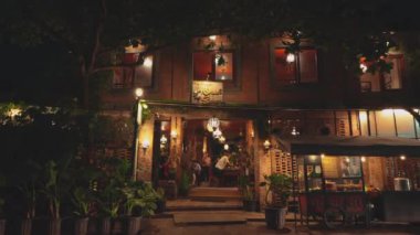 Geceleri sıcacık ışıklandırma, tuğla duvarlar ve yemyeşil yeşilliklerin olduğu büyüleyici bir restoran. Davetli girişte bir tabela ve dekoratif fenerler sergileniyor, rahat bir atmosfer yaratıyor..