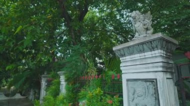 Süslü taş sütunların bulunduğu, yemyeşil ve renkli çiçeklerle çevrili sakin bir bahçe girişi. Sahne sakin ve davetkar, doğanın güzelliğini gözler önüne seriyor..