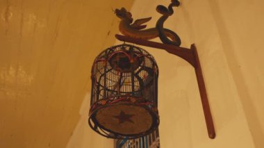 Duvardan sarkan dekoratif bir kuş kafesi. Karmaşık tasarımlar ve canlı renkler. Kafes daireseldir, tabanında bir yıldız deseni vardır, ahşap bir parantezden sarkıtılmıştır..