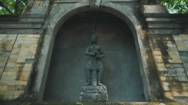 Yıpranmış taş duvarlar ve yemyeşil yeşilliklerle çevrili bir savaşçı heykeli belirgin bir şekilde kemer yolunda duruyor. Heykel ayrıntılı ve geleneksel kıyafetlerle bir figür tasvir ediyor, güç ve kültürel önem yayıyor..