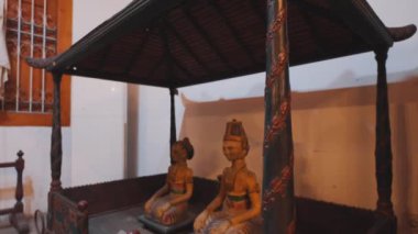 Geleneksel bir gölgelik altında oturan iki ahşap heykel, karmaşık oymalar ve canlı renkler sergiliyor. Rakamlar geleneksel kıyafetlerle süslenmiş, kültürel sanatları yansıtıyor..