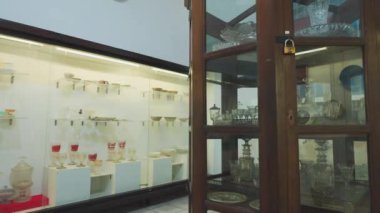 Müze ortamında çeşitli cam eşyalar ve dekoratif eşyalar sergileniyor. Cam dolapta kaseler, fincanlar ve dekoratif vazolar da dahil olmak üzere çeşitli antika ve modern cam parçaları sergileniyor..