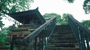 Yeşilliklerle çevrili geleneksel bir tapınak yapısına çıkan merdiven manzarası. Merdivenler taştan yapılmış ve etrafı ağaçlarla çevrili siyah bir korkuluk var..