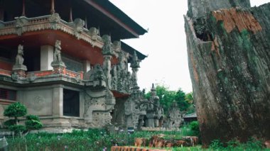 Yeşillik ve ön planda büyük, yıpranmış bir ağaç kütüğüyle çevrili karmaşık taş oymalarıyla geleneksel bir Bali tapınağı..