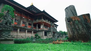 Yeşillik ve ön planda büyük, yıpranmış bir ağaç kütüğüyle çevrili karmaşık oymalar ve heykellerle dolu geleneksel bir Bali tapınağı..