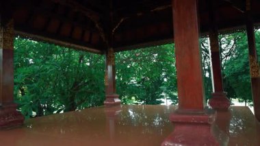 Etrafı yemyeşil ağaçlarla çevrili ahşap bir köşkten huzurlu bir manzara. Parlak zemin doğal ışığı yansıtıyor, barışçıl bir atmosfer yaratıyor..