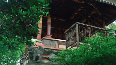 Yeşillik ve ahşap mimari ve dekoratif unsurlarla çevrili geleneksel bir Bali yapısı. Bina yüksek, süslü parmaklıklarla dolu bir balkon sergileniyor..
