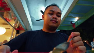 Yemek için kırmızı çubuklar kullanan, bir kase eriştenin tadını çıkaran bir adam. Şehriyeleri höpürdettiği andaki haz ifadesine odaklanmış durumda. Renkli bir yemek düzeninin bulanık arka planına sahip..