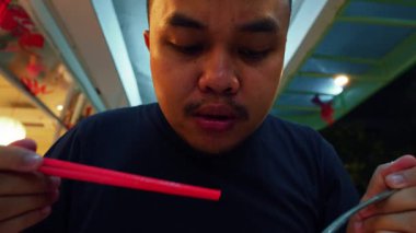 Yemeğin tadını çıkaran, kaşığı ağzına yakın tutan bir adam. Ayarlar, arka planda hafif ışıklandırma olan sıradan bir yemek alanı gibi görünüyor..