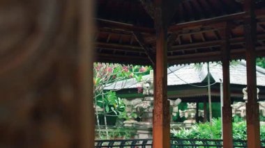 Yeşillik ve tropikal çiçeklerle çevrili geleneksel bir Bali pavyonunun huzurlu manzarası. Karmaşık ahşap mimari kültürel ayrıntıları sergiliyor, barışçıl bir atmosfer yaratıyor..