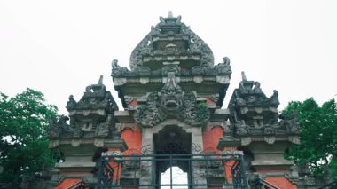 Görkemli bir Bali tapınağı kapısı. Karmaşık taş oymalarla dolu, yemyeşil bir yer. Yapıda geleneksel mimari unsurlar ve arka planda bulutlu bir gökyüzü yer alıyor..