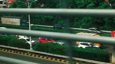 Yatay metal parmaklıklardan görünen işlek bir şehir yolu. Trafikte turuncu bir kamyon, beyaz arabalar ve kamyonetler de dahil olmak üzere çeşitli araçlar görülüyor. Yol kenarındaki yemyeşil ağaçlar kentsel ortama doğal bir kontrastlık ekliyor..