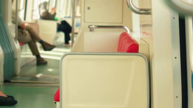 Boş kırmızı koltukları ve arka planda yolcuları olan bir metro treni manzarası. Zemin yeşil ve ışıklandırma parlak. Geniş bir atmosfer yaratıyor..