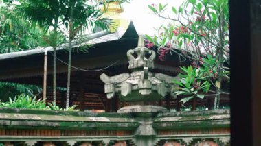Yeşillik ve tropikal bitkilerle çevrili karmaşık oymalarla süslenmiş geleneksel bir Bali mimari özelliği. Arka plan, Bali tasarımına özgü ahşap bir yapıyı gösteriyor..