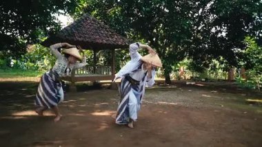 Kırsal bir ortamda geleneksel dans eden iki kişi, geleneksel kıyafetler ve hasır şapkalar giyerek, arka planda yeşilliklerle çevrili bir taraçayla.