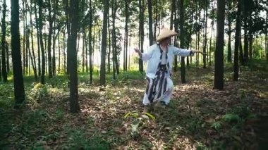 Geleneksel giyinmiş bir insan yemyeşil bir ormanda dans eder. Güneş ışığı ağaçların arasından süzülerek sakin bir atmosfer yaratır. Bireyler neşeli ve anlamlı görünürler, doğayla çevrili..