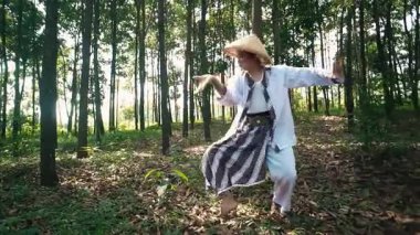 Geleneksel konik şapka ve beyaz giysi giyen bir kişi ormanda duruyor ve ok atmaya hazırlanıyor. Sahne uzun ağaçlar ve düşen yapraklarla çevrili, sakin bir doğal ortam yaratıyor..