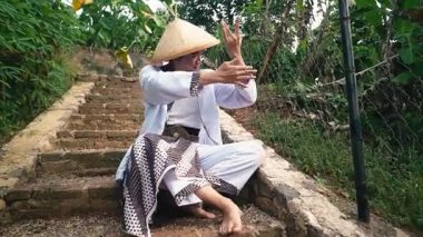 Geleneksel konik şapka ve kültürel kıyafet giyen bir insan yeşil alanlarla çevrili taş basamaklarda dans eder. Dansçının elleri zarif bir pozisyonda, zarif bir hareket sergiliyor..