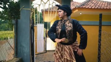 Geleneksel giyinmiş bir kişi, arka planda yeşil tarlaları ve canlı sarı bir evi olan bir kapıda güvenle duruyor..