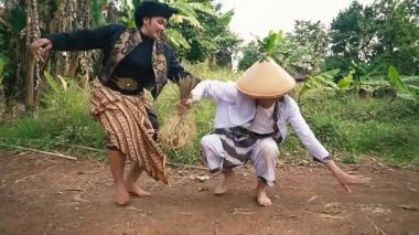 Geleneksel giyinmiş iki kişi, arka planda muz ağaçları olan kırsal bir ortamda eğlenceli bir kavgaya tutuşmuş..