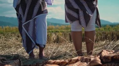 Kültürel giysiler içinde tarlada dikilen geleneksel eşyaları tutan iki birey. Arka planda dağlar ve mavi gökyüzüyle manzaralı bir manzara var..