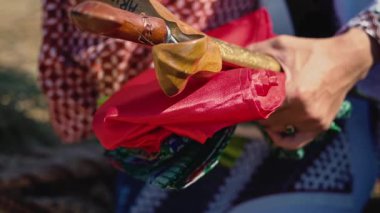 Kırmızı, yeşil ve desenli kumaşlar da dahil olmak üzere renkli kumaşları tutan bir kişinin yakın plan fotoğrafı, diğer elde tahta bir sopa veya baston, doğal bir arka plana karşı dizilmiş..