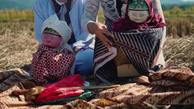 Geleneksel bir sahnede iki kişi tarlada desenli bir kumaşa oturmuş, ellerinde geleneksel kıyafetlerle renkli bebekler tutuyorlardı. Bebeklerin farklı yüz hatları var ve kültürel eserlerle çevrili dekoratif kumaşlara sarılı..