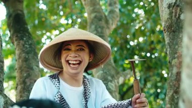 Geleneksel konik şapka takan genç bir kadın, yemyeşil ağaçların arasında otururken gülümsüyor. Sahne neşe ve kültür mirasını aktarıyor.