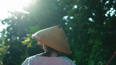 Geleneksel konik şapka takan bir kişi, ağaçların arasından süzülen güneş ışığıyla yemyeşil bir ortamda duruyor. Odak noktası figürün arkası. Huzurlu ve huzurlu bir atmosfer yaratıyor..