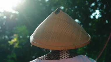 Geleneksel konik şapka takan bir insan, ağaçların arasından süzülen güneş ışığıyla yemyeşil bir ortamda yürür..