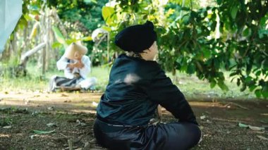 Bereketli yeşil bir çevrede iki kişinin olduğu sakin bir açık hava sahnesi. Bir kişi yerde oturuyor, siyah giyinmiş, diğeri ise arka planda, derin düşüncelere dalmış bir şekilde.. 