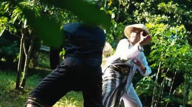 İki dövüş sanatçısı açık havada yemyeşil bir mekanda dinamik bir dövüş sahnesine çıktılar. Biri konik şapkalı geleneksel bir kıyafet giyer, diğeri ise modern giysiler içinde, bir tarz karışımı sergiler..