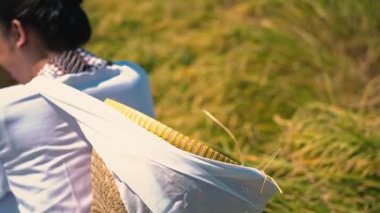 Geleneksel giyimli bir kadın pirinç tarlasında otururken arkadan görülüyor. Sırtında örülmüş bir sepet taşıyor ve büyük bir hasır şapka tutuyor. Altın pirinç bitkileri onu çevrelemiş, dingin bir tarım manzarası yaratmış..