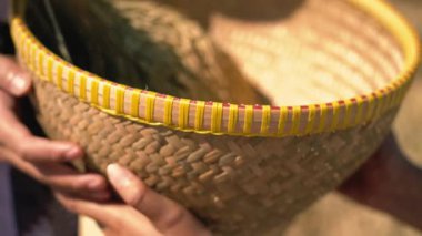 Ellerin tuttuğu, karmaşık desenler ve sarı süslemeli dokuma sepetinin yakın çekimi. Sepet, bulanık doğal bir arka plana kurulmuş kuru ot ya da saman içeriyor..