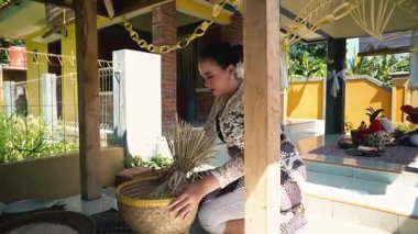 Geleneksel giysili bir kadın dokuma sepetinin yanında diz çöküp dışarıda dekoratif eşyalar düzenliyor. Arka planda renkli bir ev ve yemyeşil bir ortam var..