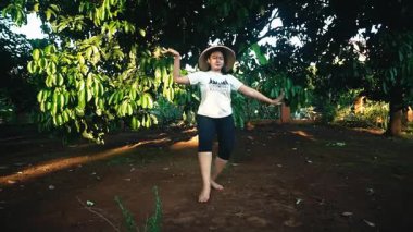 Yeşil yemyeşil bir bahçede saman şapka ve günlük giysiler içinde neşeyle dans eden bir insan. Güneş ışığı ağaçların arasından süzülüp sıcak bir atmosfer yaratır..