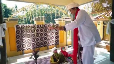 Geleneksel Endonezya dekoratif kumaşlar ve geleneksel giysiler giymiş iki oyuncakla beyaz elbiseli bir kişinin yer aldığı geleneksel bir Endonezya seti. Sahne parlak ve kültürel öğeler sergiliyor..