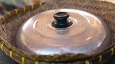 Dokuma sepetinin üzerinde duran metalik bir kapağın yakın çekimi, kırsal bir mutfak ortamı sergiliyor. Kapağın parlak bir yüzeyi ve siyah bir topuzu var. Sepet de karmaşık bir şekilde sarı aksanla örülmüş..