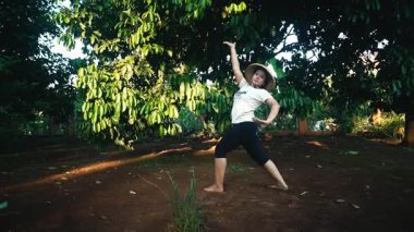 Yeşillik yeşillik ve ağaçlarla çevrili doğal bir ortamda dans pozu veren bir insan. Kişi hasır şapka ve sıradan bir tişört giyer. Zarif bir hareket sergiler..