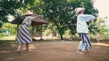 Geleneksel Endonezya kıyafeti giymiş iki çocuk kırsal bir açık hava ortamında oynuyorlar. Bir çocuk beyaz bir gömlek ve desenli bir sarong giyerken, diğeri benzer bir kıyafet giyiyor..