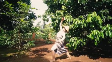 Yeşil bitki ve ağaçlarla çevrili yemyeşil bir bahçede diz çökmüş, konik şapkalı bir kadın. Dışarıdaki bir etkinlikte kıyafetlerini düzeltiyor gibi görünüyor..